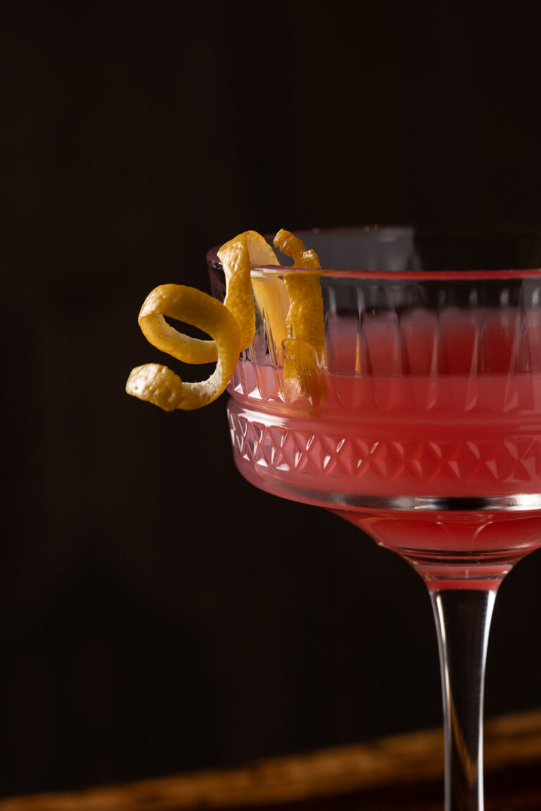 A pink cocktail in a martini glass with an orange peel garnish.