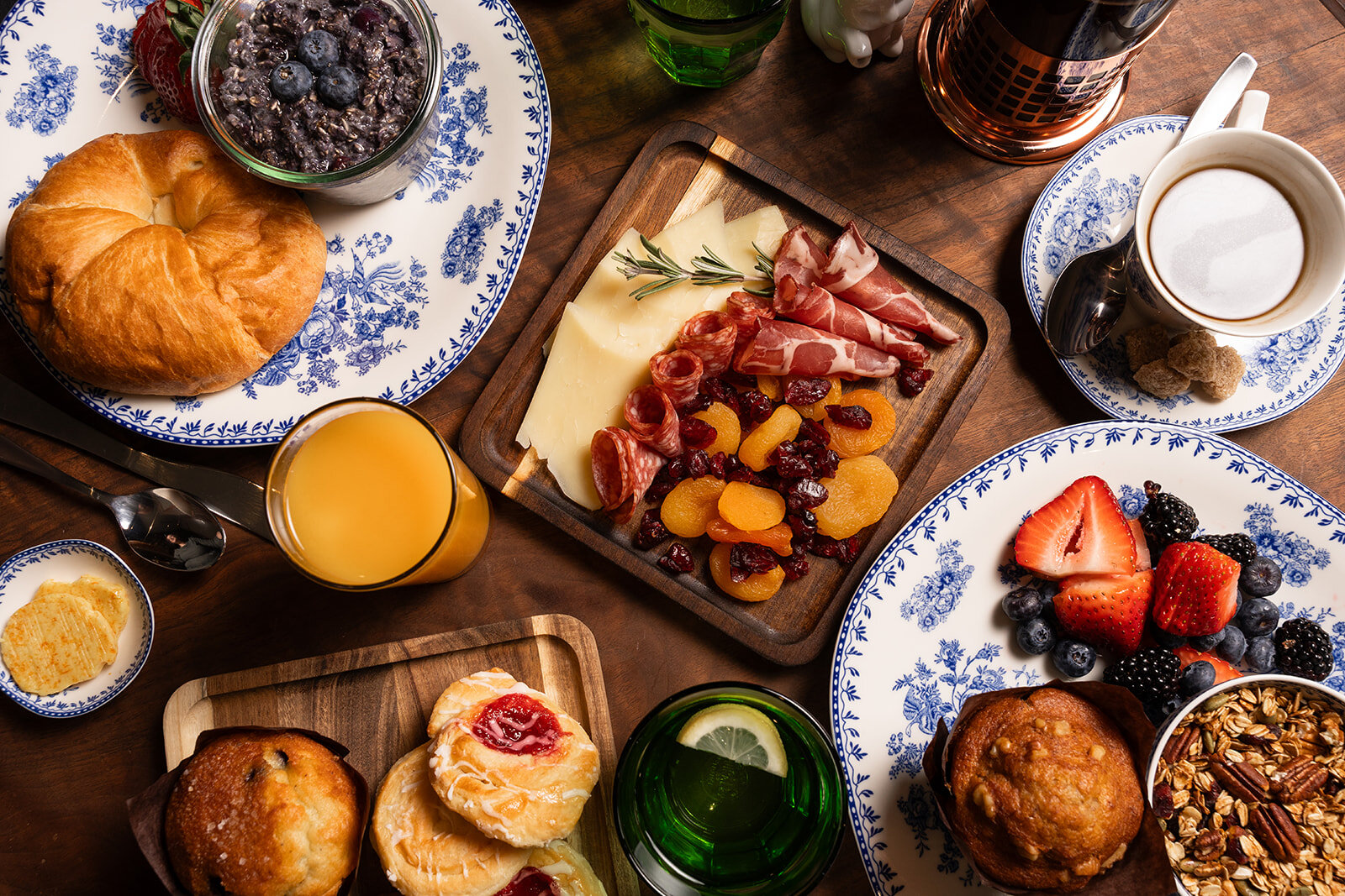 A breakfast spread on a wooden table with various pastries, fruits, cheese, and charcuterie