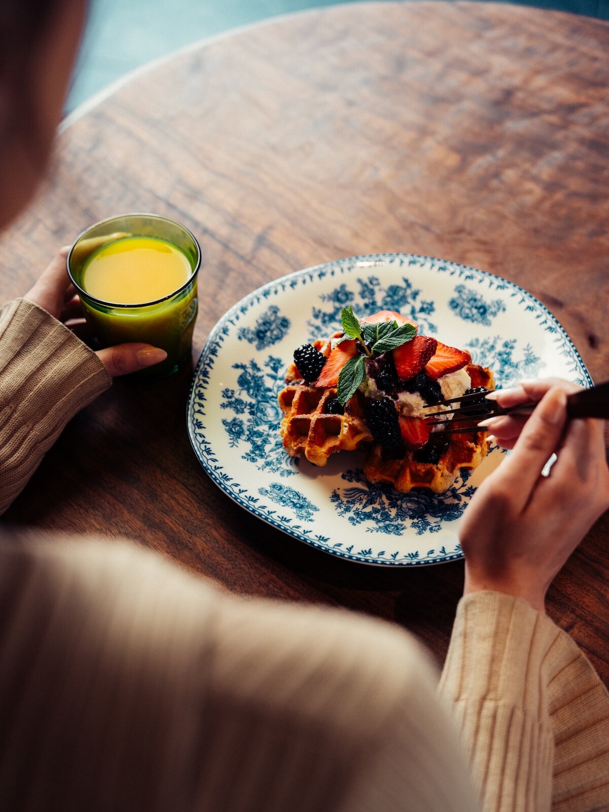 A person holding a glass of orange juice and a fork with a waffle topped with strawberries, blackberries, and mint.